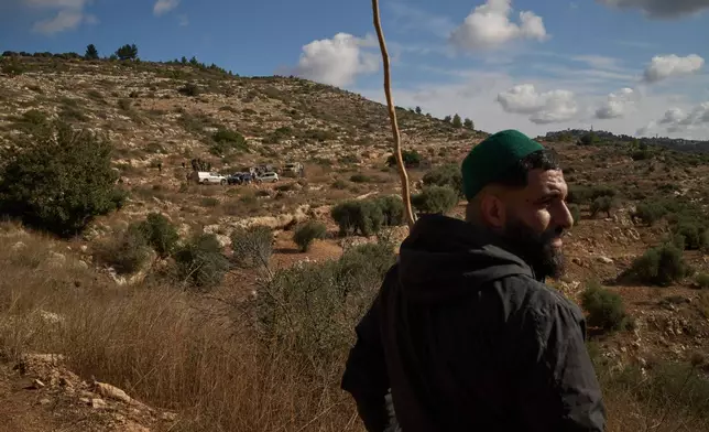 A Palestinian looks on after being prevented from continuing to harvest olives by Israeli settlers and soldiers, who stand next to vehicles in the background, in the West Bank village of Deir Abu Mashal, Monday, Oct. 19, 2026. (AP Photo/Leo Correa)