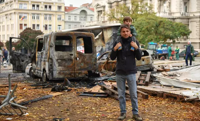 A man and child pose by burnt-out vehicles in a park used as a camp by supporters of the right-wing Serbian Progressive Party (SNS) where tents and vehicles were set on fire by celebrating crowds after Aleksandar Vucic, the party leader conceded defeat in the parliamentary election, held on Sunday, in Belgrade, Serbia, Monday, Oct. 26, 2026. (AP Photo/Armin Durgut)