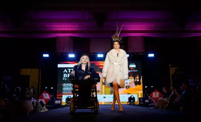 Andreea Lichi, a wheelchair user, is accompanied on the catwalk by a model during the Atypic Beauty Gala fashion show, an event promoting inclusion and equal opportunities for people with disabilities, in Bucharest, Romania, Thursday, Oct. 22, 2026. (AP Photo/Andreea Alexandru)