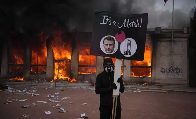 A protester holds a board with a photo of French president Emmanuel Macron in front of a burning tourism office during a demonstration against rising living costs and in solidarity with students’ grievances in Lyon, France, Saturday, Oct. 17, 2026. (AP Photo/Laurent Cipriani)