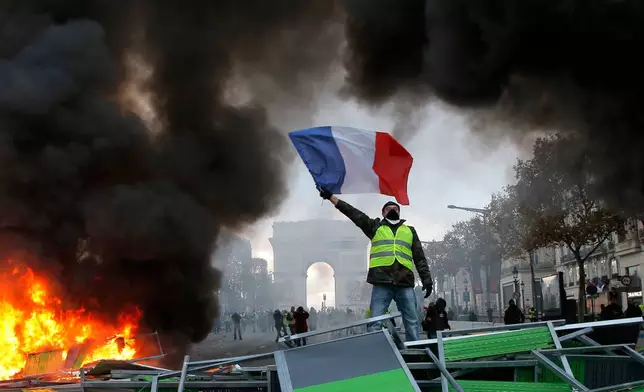 FILE - A demonstrator wearing a yellow vest waves the French flag on top of a burning barricade on the Champs-Elysees avenue with the Arc de Triomphe in background in Paris, on Saturday, Nov. 24, 2018, during a demonstration against the rising fuel taxes. (AP Photo/Michel Euler, File)