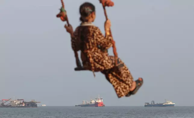 A girl swings along the shore as cargo ships and other commercial vessels appear anchored in the Strait of Hormuz off Bandar Abbas, Iran, Tuesday, Oct. 13, 2026. (Razieh Poudat/ISNA via AP)