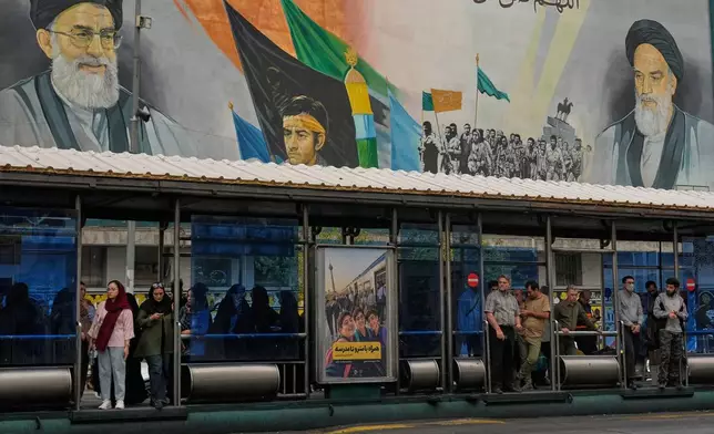 Commuters stand at a bus stop on Islamic Revolution street in front of a mural depicting the late Iranian revolutionary founder Ayatollah Khomeini, right, the late Supreme Leader Ayatollah Ali Khamenei, who was killed in the U.S. and Israel strike on Feb. 28, left, and Basij paramilitary forces, in Tehran, Iran, Tuesday, Oct. 6, 2026. (AP Photo/Vahid Salemi)