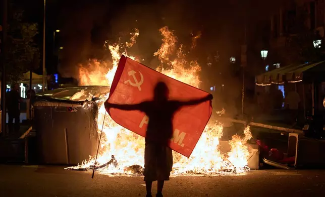 A protesters holds a flag next to trash cans on fire during clashes with police following a demonstration over Spain's housing crisis, hours after Prime Minister Pedro Sánchez announced an early general election, in Barcelona, Spain, Monday, Oct. 5, 2026. (AP Photo/Emilio Morenatti)
