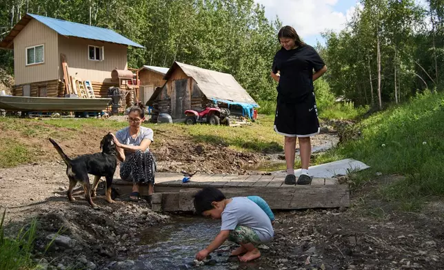 Lewis Thomas, front, plays in a creek as his cousin Alara Woods, right, watches and his mother, Brooke Woods, shovels gravel at their home in Rampart, Alaska, Saturday, July 11, 2026. (AP Photo/Annika Hammerschlag)