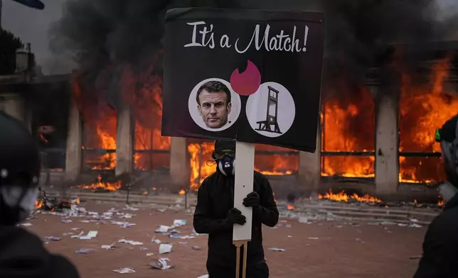 A protester holds a placard with a photo of French president Emmanuel Macron and a guillotine in front of a burning tourism office during a demonstration against rising living costs and in solidarity with students' grievances in Lyon, France, Saturday, Oct. 17, 2026. (AP Photo/Laurent Cipriani)
