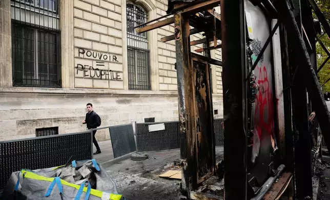 A man walks past a burned-out structure next to the inscription in a wall ¨Power = Pedophilia¨ after the weekend protests over the cost of living turned into rioting and looting across France, in Paris, Wednesday, Oct. 21, 2026. (AP Photo/Teresa Suarez)