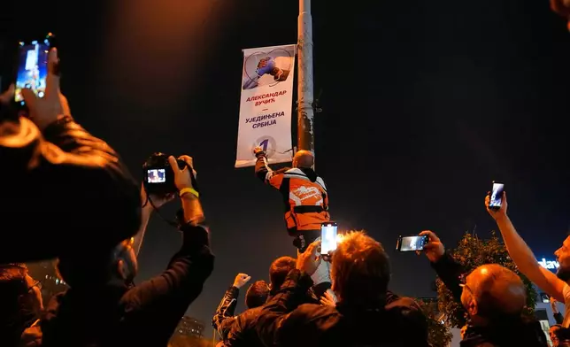 People shoot photos and video on their mobile phones as a man tears down a poster of Aleksandar Vucic's, right-wing Serbian Progressive Party (SNS) after he conceded defeat in the parliamentary election in Belgrade, Serbia, Sunday, Oct. 25, 2026. (AP Photo/Petr David Josek)