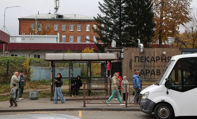 People are seen at a bus stop in Shelekhov outside Irkutsk, Russia, on Thursday, Oct. 8, 2026, backdropped by the district hospital and the medical center for treating infectious diseases. (AP Photo)