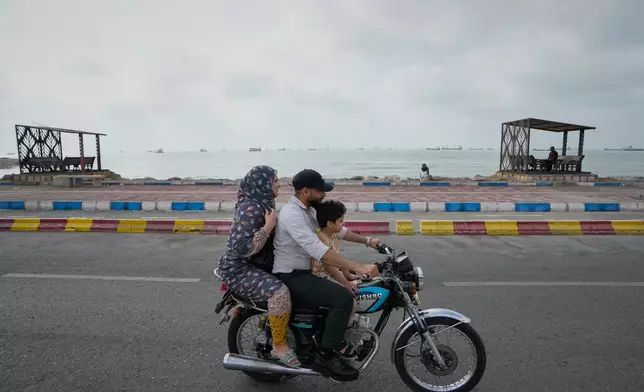 一名男子、女子及男童沿海濱騎電單車，商船停泊在伊朗班達阿巴斯對開的霍爾木茲海峽。攝於2026年9月4日周五。(美聯社圖片/Vahid Salemi) AP圖片